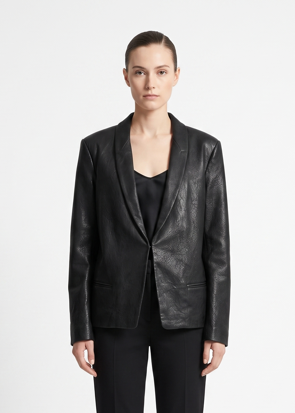 A model wearing a black textured leather blazer with shawl lapels over a black top and black trousers, photographed against a plain light gray studio background.