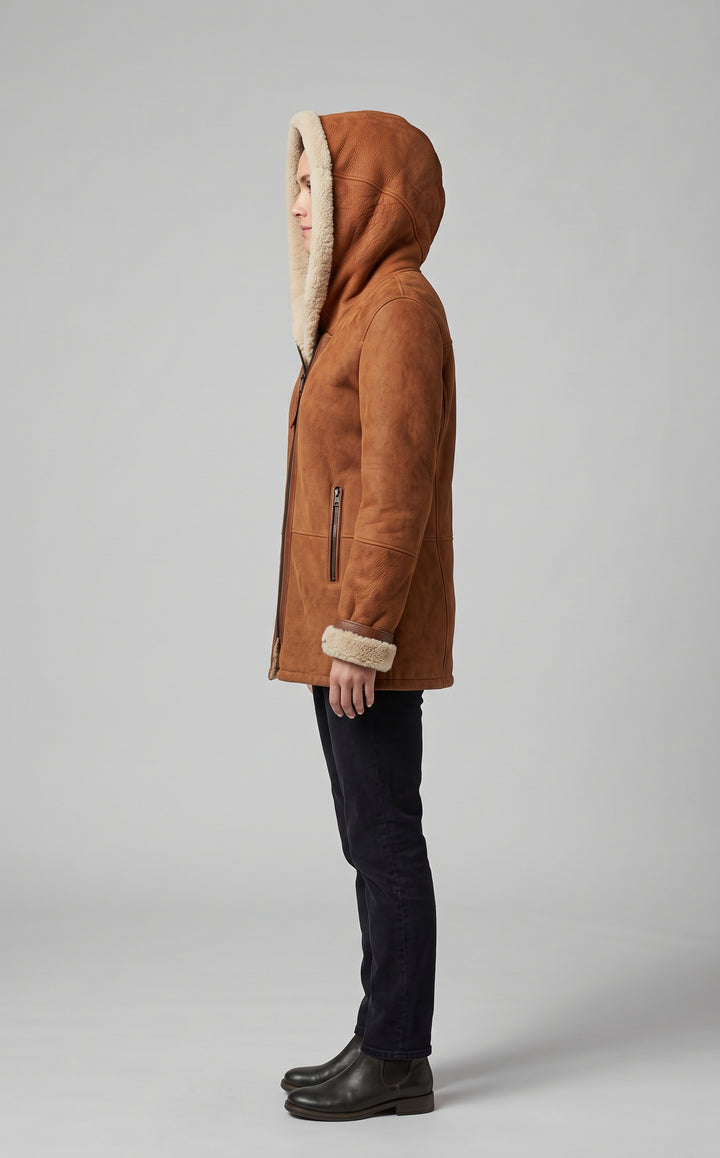 A side-profile view of a person wearing a brown suede hooded jacket with a cream-colored shearling lining, paired with dark jeans and black boots, against a neutral gray studio background.