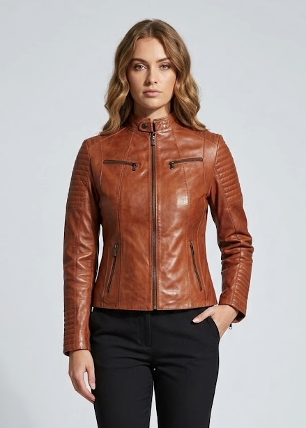 A woman modeling a fitted brown leather moto jacket with a stand collar, zippered front, and zip chest pockets, paired with black trousers and black ankle boots, against a light gray studio background.
